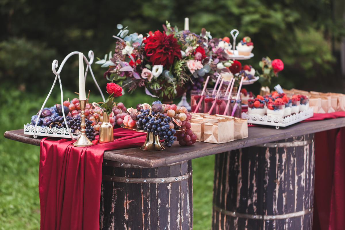 Wedding reception wooden table standing on two barrels, decorated with red flowers, sweets, drinks and silk. Rustic style
